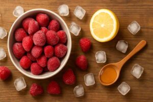 Top-down view of frozen strawberries spilling from a bowl, with ice cubes, lemon halves, and a spoonful of honey on a wooden table, highlighting fresh smoothie ingredients.